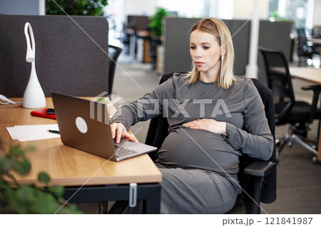 A pregnant woman sits at a desk in the office, with some personal items on the table, laptop and a plant, in a typical white collar worker setup A pregnant woman sits at a desk in the office, with some personal items on the table, laptop and a plant, in a typical white collar worker setup 121841987