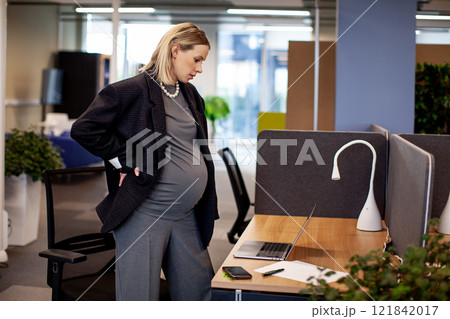 Pregnant woman standing in office cubicle, surrounded by a desk, computer, chair, and office supplies. Portrait of professional pregnant businesswoman at work 121842017