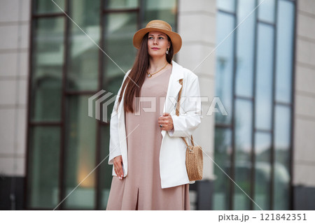 Stylish Plus size woman Standing outdoors in urban city, wearing beige dress, white blazer, wide brimmed hat, woven handbag. Model with long brown hair Stylish Plus size woman Standing outdoors in urban city, wearing beige dress, white blazer, wide brimmed hat, woven handbag. Model with long brown hair 121842351