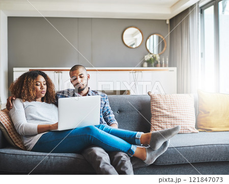Relaxed couple relaxing at home using an online streaming service on a laptop and enjoying the day together. Young African American lovers sitting on a couch resting and watching an internet video Relaxed couple relaxing at home using an online streaming service on a laptop and enjoying the day together. Young African American lovers sitting on a couch resting and watching an internet video 121847073