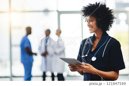Young smiling medical professional doctor, holding digital tablet in modern hospital. Female healthcare specialist standing with test results, with colleagues in background. 121847589