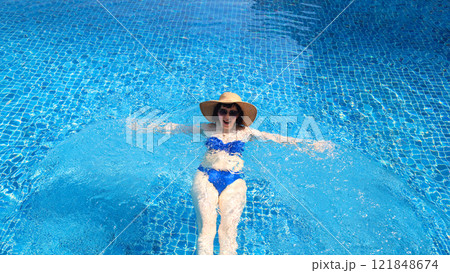 Blissful Summer Escape: A Woman in a Straw Hat and Sunglasses Joyfully Swimming in a Private Pool, Embracing the Relaxation and Serenity of a Sun-Kissed Day Under the Clear Blue Sky 121848674