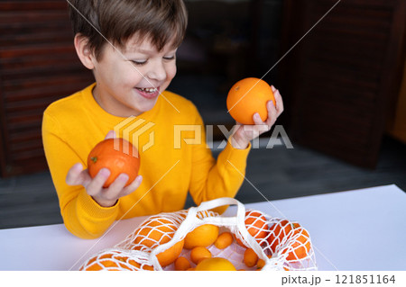 A child with a smile holds an orange and a mandarin, looking at a bag filled with citrus fruits.  121851164