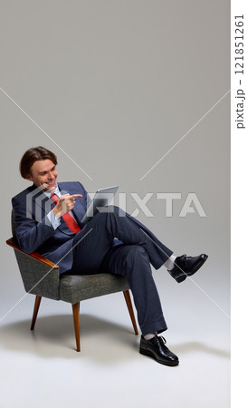 Man sitting in chair, dressed suit with red tie, holding tablet and pointing at screen with cheerful expression against gray studio background. 121851261