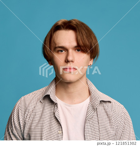 Young man wearing plaid shirt over white t-shirt, looking directly at camera with neutral expression, standing against plain blue studio background. Young man wearing plaid shirt over white t-shirt, looking directly at camera with neutral expression, standing against plain blue studio background. 121851302