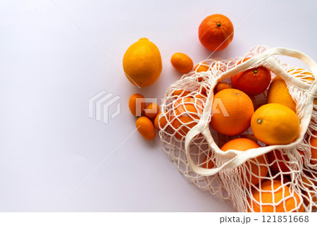 Bright citrus fruits in a string bag on a white background, with a few fruits lying nearby. 121851668