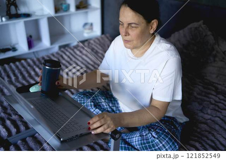 a senior woman works from home on a laptop and drinks coffee from a thermos mug a senior woman works from home on a laptop and drinks coffee from a thermos mug 121852549