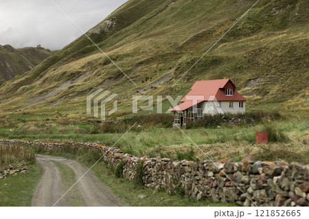 A charming house with a red roof rests by a winding dirt road in the Georgian autumn hills, surrounded by rolling slopes and a cloudy daytime sky 121852665