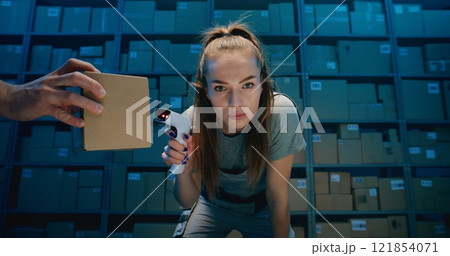 Tired Female Warehouse Employee Looking at Camera, Scanning Cardboard Boxes Using Scanner 121854071
