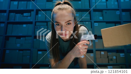 Tired Female Logistics Specialist Looking at Camera, Scanning Cardboard Boxes with Scanner in Warehouse 121854101