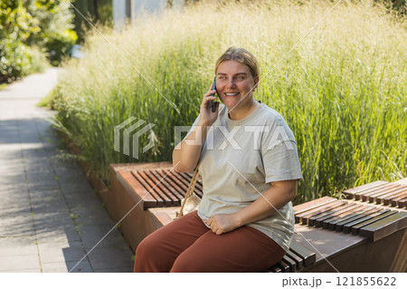 30s cheerful woman dressed in casual look sitting on the bench and talking on smartphone on the city street. Female relaxing outdoors. Urban concept. 30s cheerful woman dressed in casual look sitting on the bench and talking on smartphone on the city street. Female relaxing outdoors. Urban concept. 121855622