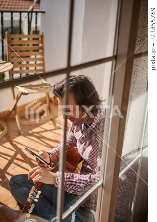 Man sitting outside with guitar checking smartphone on a sunny terrace 121855789