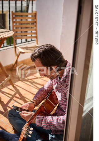 Man playing guitar and checking his phone on a sunny balcony Man playing guitar and checking his phone on a sunny balcony 121855790