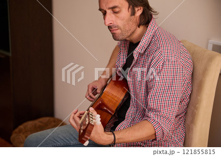Man playing an acoustic guitar while seated indoors in a casual setting 121855815