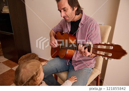 Father playing guitar for his young child in a cozy living room 121855842