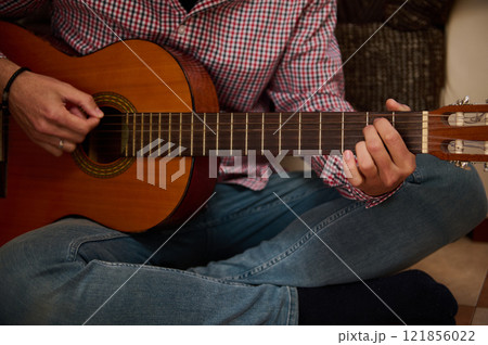 Person playing acoustic guitar while seated on the floor at home 121856022