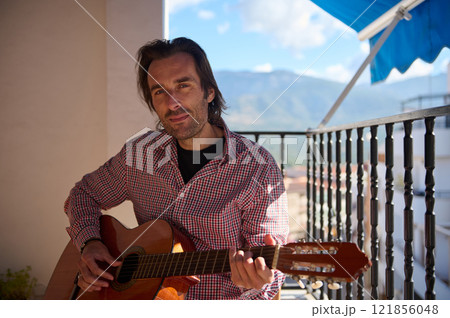 Man playing a guitar on a balcony with a scenic mountain view 121856048