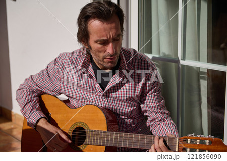 Man playing an acoustic guitar outdoors near a window in natural light Man playing an acoustic guitar outdoors near a window in natural light 121856090