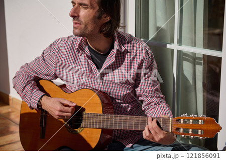 Man playing guitar outdoors while seated in sunlight near a window Man playing guitar outdoors while seated in sunlight near a window 121856091