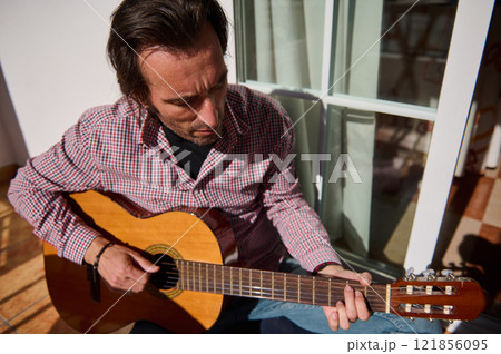 Person playing acoustic guitar outdoors on a sunny day near a window 121856095