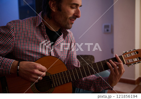 Man seated and playing acoustic guitar in a cozy indoor setting Man seated and playing acoustic guitar in a cozy indoor setting 121856194