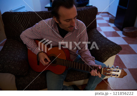 Man playing a guitar while sitting on a couch in a cozy living room Man playing a guitar while sitting on a couch in a cozy living room 121856222
