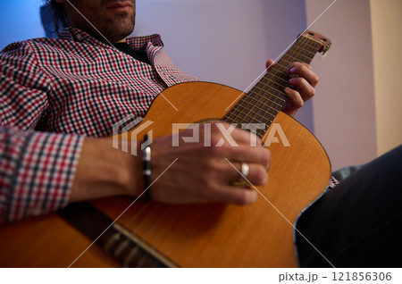 Person sitting and strumming an acoustic guitar indoors wearing casual clothing Person sitting and strumming an acoustic guitar indoors wearing casual clothing 121856306