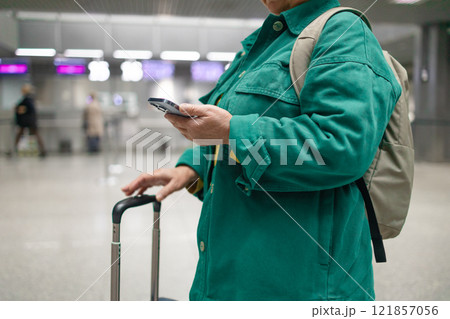 Caucasian 60s smiling business woman in green jacket in international airport, checking her phone while waiting for her flight 121857056