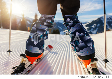 Scenic back POV view kid beginner skier close-up legs downhill start line rows freshly prepared groomed ski slope piste bright warm morning sunrise sky background.  mountain landscape winter resort 121857211