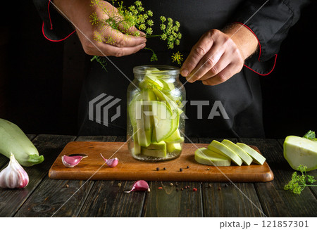 Hands skillfully arrange fresh garlic and dill on top of sliced cucumbers inside a jar. A wooden board holds additional cucumber slices, showcasing the art of preserving flavors 121857301