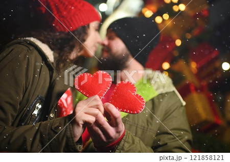 a couple of lovers - a man and a woman kiss and hold in their hands two red hearts made of felt against the background of snow and gifts 121858121