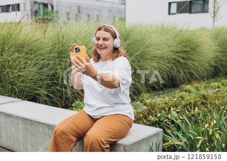 Portrait of young happy woman walking on the street in the city. Girl talking on a video call with phone and headphones outdoor, blogger records stories. Online communication concept. 121859158
