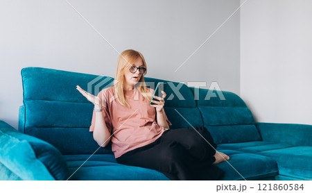 Upset sad, skeptical, serious woman holding smartphone gadget for communicate indoors. Negative people emotion, pensive girl sitting on sofa at home. 30s woman holding smartphone. 121860584