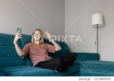 Hi. Female in taking a selfie photo using her mobile phone at home. People lounge concept. 30s girl showing peace sign with fingers. Full Body 121860585