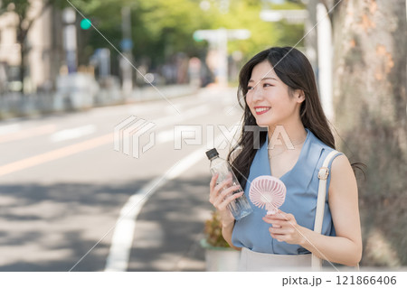 ハンディファンとペットボトルの水を持って木陰で涼むビジネスウーマン 121866406