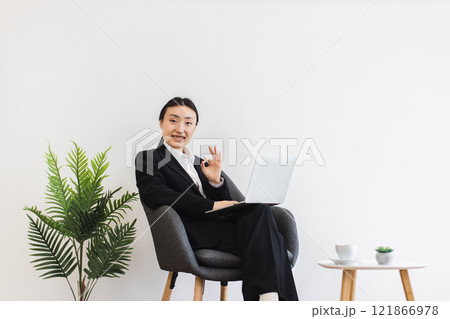 Asian businesswoman in her 30s sitting in chair using laptop with positive expression, modern office environment, potted plant and coffee table creating professional yet comfortable atmosphere. 121866978
