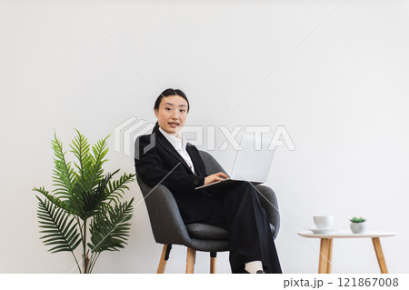 Asian businesswoman sitting on chair using laptop in modern office. Young adult female wearing business suit. Minimalist workspace design with natural plant decor suggests professional environment. Asian businesswoman sitting on chair using laptop in modern office. Young adult female wearing business suit. Minimalist workspace design with natural plant decor suggests professional environment. 121867008