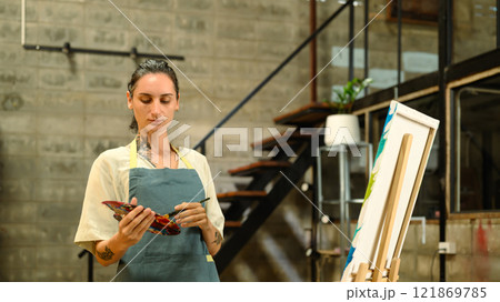 Focused Caucasian artist in an apron holding paint palette working on her art in the industrial style studio Focused Caucasian artist in an apron holding paint palette working on her art in the industrial style studio 121869785