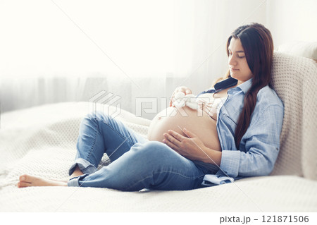 A pregnant woman is seen resting comfortably on bed, holding a pair of baby shoes near her belly. She appears content and serene, enjoying a quiet moment A pregnant woman is seen resting comfortably on bed, holding a pair of baby shoes near her belly. She appears content and serene, enjoying a quiet moment 121871506