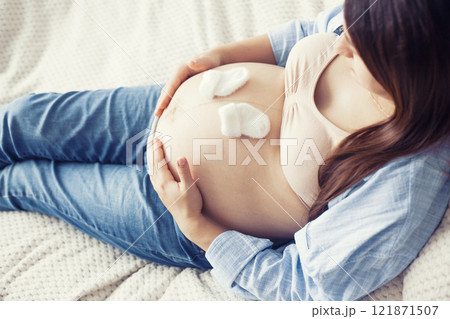 A pregnant woman is seen resting comfortably on bed, holding a pair of baby shoes near her belly. She appears content and serene, enjoying a quiet moment A pregnant woman is seen resting comfortably on bed, holding a pair of baby shoes near her belly. She appears content and serene, enjoying a quiet moment 121871507