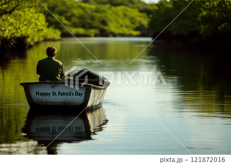 A man is rowing a small boat on a calm river surrounded by green trees, with "Happy St. Patrick's Day" on the boat. Concept of solitude and nature. 121872016