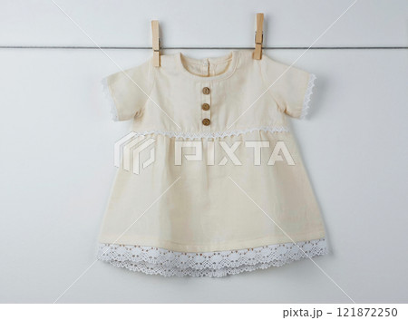 Beige baby dress with lace accents and wooden buttons hanging on a clothesline, showcasing a vintage-inspired handmade design Beige baby dress with lace accents and wooden buttons hanging on a clothesline, showcasing a vintage-inspired handmade design 121872250