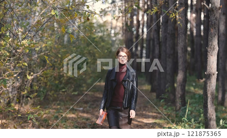 Smiling city woman relaxing in the forest. Thoughtful woman in glasses and office clothes walking in the autumn park, breathing fresh air, enjoying nature. 121875365