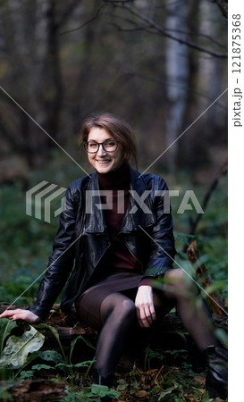 Smiling city woman relaxing in the forest. Thoughtful woman in glasses and office clothes sitting casually on an old fallen tree covered with moss outdoors, breathing fresh air, enjoying nature. Smiling city woman relaxing in the forest. Thoughtful woman in glasses and office clothes sitting casually on an old fallen tree covered with moss outdoors, breathing fresh air, enjoying nature. 121875368