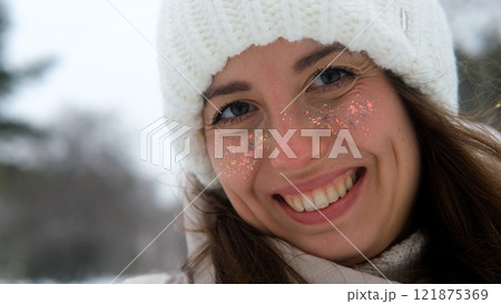 Beautiful woman with long brown hair, in warm winter clothes, standing on a snowy street in winter. She smiles and enjoys the snow, laughs and looks at the camera. Close-up 121875369