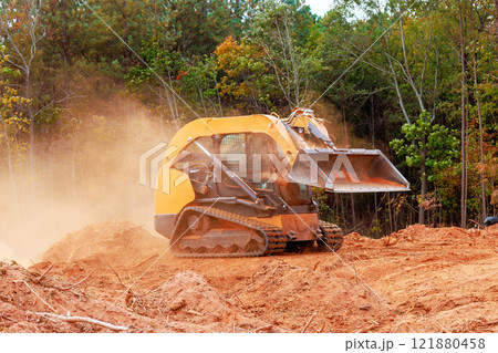 Skid steer loader works on clearing earth amid trees, creating dusty environment during preparing land for development Skid steer loader works on clearing earth amid trees, creating dusty environment during preparing land for development 121880458