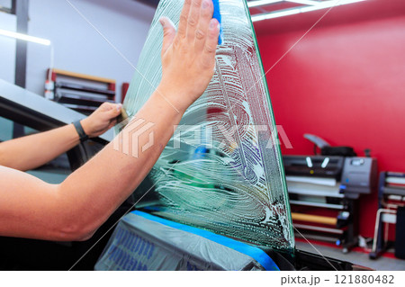 Worker is carefully applying window tint film to car rear window in a well lit workshop setting. 121880482