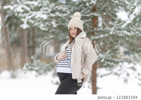 Young pregnant woman in a snowy winter forest looking at the camera with copy space. Expecting mother smiling, looking at her belly outdoors, natural light. Concept of maternity and joy of pregnancy 121881504