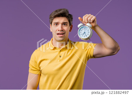 Woken up by alarm clock sleepy handsome man holding it in hand.violet background 121882497