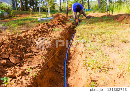 Construction worker is laying down blue PVC water pipe in freshly dug trench for irrigation in garden surrounded by trees. Construction worker is laying down blue PVC water pipe in freshly dug trench for irrigation in garden surrounded by trees. 121882738